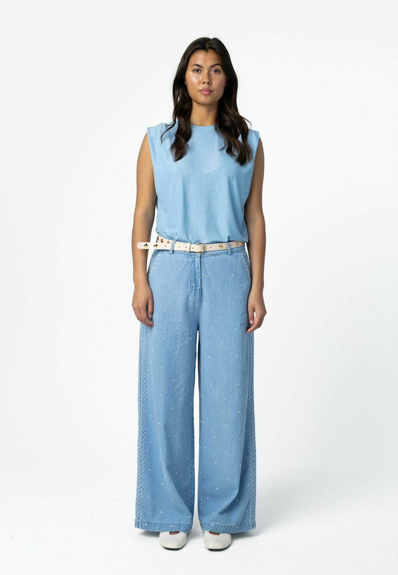 Femme debout portant un haut sans manches bleu clair, un pantalon large bleu clair brodé avec ceinture blanche et des chaussures blanches sur fond blanc.
