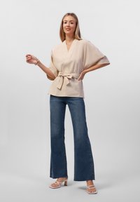 Blusa avvolgente beige a maniche corte, con vita a vita alta, abbinata a jeans blu a vita alta e zeppe bianche con cinturini. Tessuto dalla trama liscia.