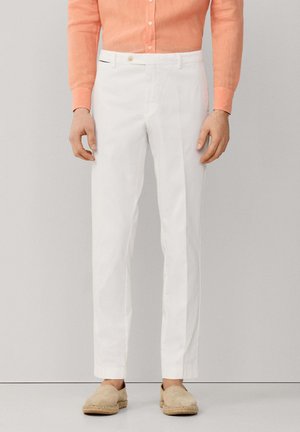 Hombre con blazer marrón oscuro, camisa coral abotonada, pantalones blancos, zapatos beige sin cordones y un pañuelo de bolsillo azul con estampado, de pie contra una pared gris.