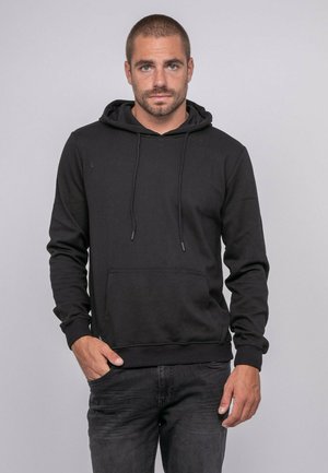 Homme aux cheveux courts et à la barbe, portant un sweat à capuche noir et un jean sombre, debout avec une main dans la poche, sur un fond gris uni.