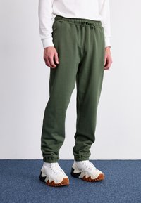 Pantalones de chándal verdes con cintura y puños elásticos, con bolsillos con cremallera y una textura suave, combinados con zapatillas blancas y negras.