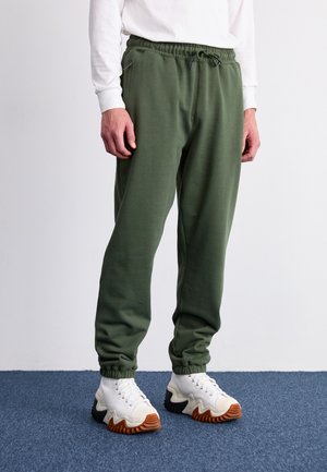 Pantalones de chándal verdes con cintura y puños elásticos, con bolsillos con cremallera y una textura suave, combinados con zapatillas blancas y negras.