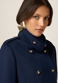 Marineblauer Wollmantel mit hohem Kragen, ausgestattet mit goldenen Knöpfen und einem strukturierten Stoff. Große, glänzende goldene Ohrringe betonen das Outfit.