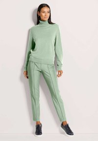 Frau in einem hellgrünen Rollkragenpullover und passenden Hosen, die vor einem hellen Hintergrund steht und schwarze Turnschuhe trägt.