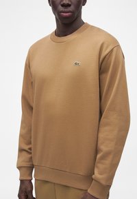 Homme portant un sweat-shirt à manches longues marron clair avec un petit logo crocodile vert sur le côté gauche de la poitrine.