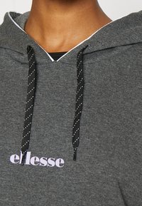 Harmaa huppari, jossa on V-kaulus, mustavalkoiset raidalliset nauhat ja rintakehällä violetti "ellesse"-logo.