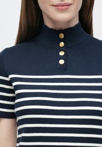 Marineblaues, gestricktes Polooberteil mit weißen horizontalen Streifen, einem hohen Kragen und fünf goldfarbenen Knöpfen auf der Knopfleiste.