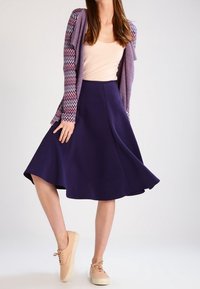 Fioletowa rozkloszowana spódnica z gładkiego materiału, zestawiona z jasnym topem i beżowymi trampkami. Strój uzupełnia wzorzysty kardigan.