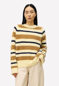 Vrouw in een fuzzy gestreepte trui in crème, bruin en marineblauw, gecombineerd met hoog-waisted lichtbeige broek, staand met één hand in de zak.