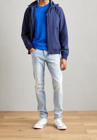 Granatowa kurtka na suwak z kapturem, niebieski t-shirt z okrągłym dekoltem, jasne jeansy oraz białe trampki na drewnianej podłodze.