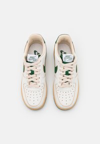 Paire de baskets en cuir blanc Nike Air Force 1 avec logos swoosh verts et lacets beiges vues de dessus sur fond blanc.