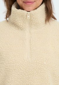 Kremowy polarowy polar z wysokim kołnierzem, zamkiem błyskawicznym i fakturowaną powierzchnią. Dyskretne logo wyszyte na piersi.