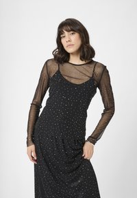 Donna con capelli castani di lunghezza spalla che indossa un abito nero con maniche lunghe trasparenti e pois argentati, in posa su uno sfondo bianco.