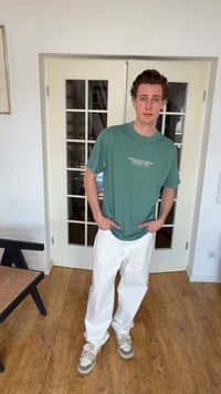 Jeune homme debout à l'intérieur, les mains dans les poches, portant un t-shirt graphique vert, un pantalon blanc et des baskets, devant des portes vitrées.