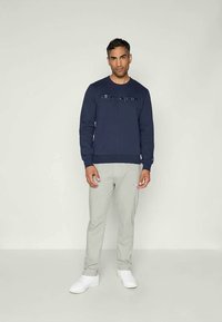 Felpa blu navy con logo, polsini e orlo a costine; abbinata a pantaloni grigi chiari con un piccolo logo e sneakers bianche.