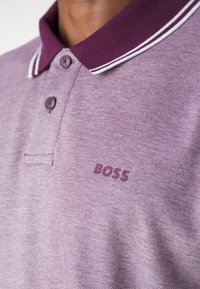 Polo de couleur violet clair avec un motif texturé, doté d'un col violet foncé avec des rayures blanches et d'un logo "BOSS" sur la poitrine.