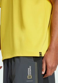 Gul t-skjorte med en glatt tekstur, med et National Geographic-logo merke på kanten. Grå shorts med gule detaljer og logo synlig.