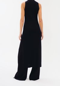 Abito nero a coste con collo alto, design senza maniche e spacco laterale, indossato su pantaloni neri a gamba larga. Texture liscia.