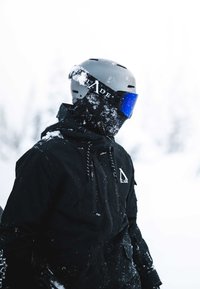 Black snow jacket with drawstrings and logo; grey ski helmet with blue reflective visor; snow on jacket and helmet; snowy background.