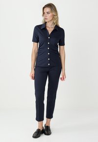Marineblauwe blouse met korte mouwen en een kraag, voorzien van gouden knopen, gecombineerd met aansluitende marineblauwe broek en zwarte loafers. Zichtbare gladde stoftextuur.