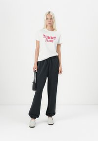 Balta marškinėliai su raudonu užrašu "TOMMY JEANS", derinami su juodais sportiniais kelnais ir šviesiais bateliais su užsegimu. Modelis laiko mažą juodą rankinę.