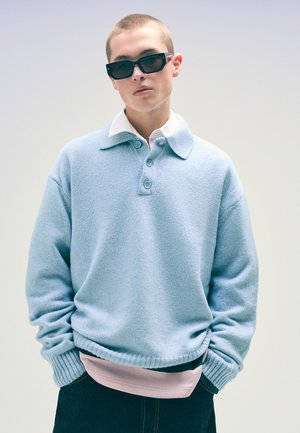 Lys blå fleece-pullover med knaplukning ved kraven, ribbede manchetter og en lagdelt pink skjorte underneath. Sorte solbriller fuldender looket.