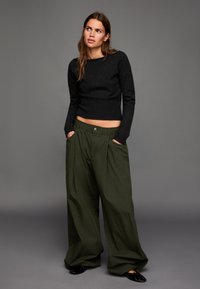 Fekete kötött pulóver hosszú ujjal és bordázott szegéllyel, oversized sötétzöld nadrággal párosítva, amelyen gombos záródás és oldalzsebek találhatók.