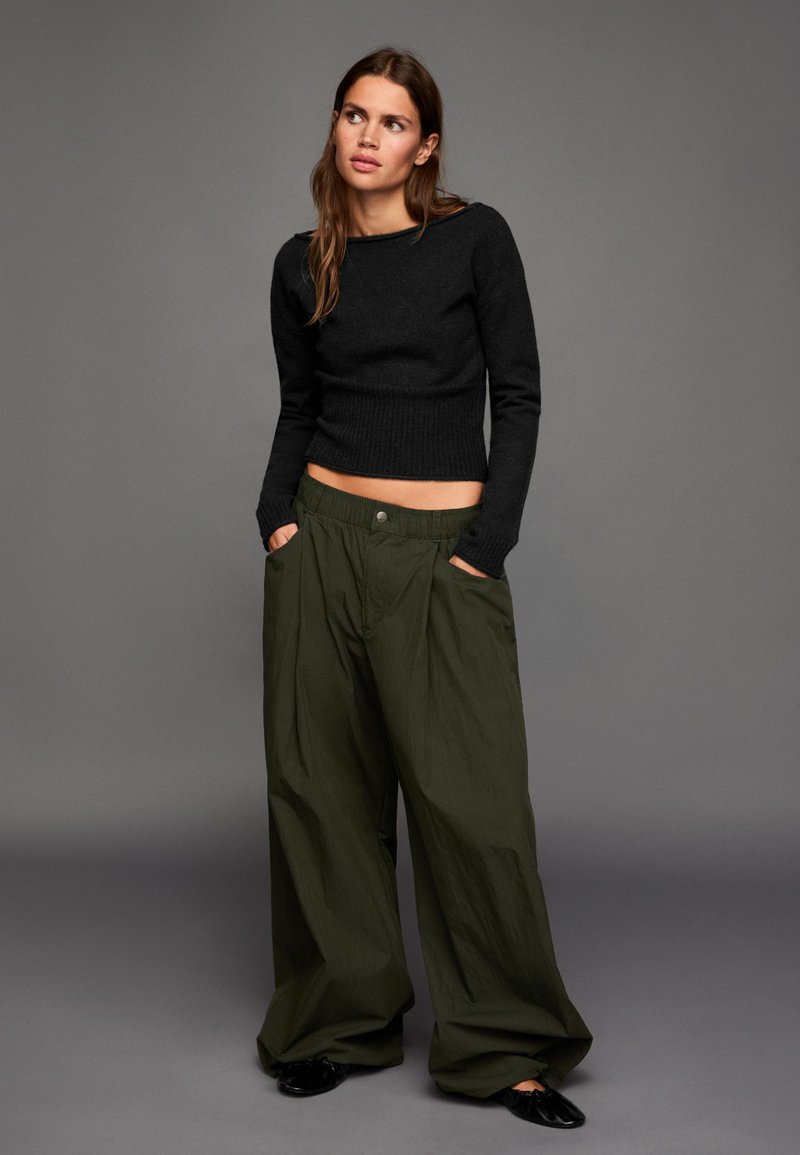 Fekete kötött pulóver hosszú ujjal és bordázott szegéllyel, oversized sötétzöld nadrággal párosítva, amelyen gombos záródás és oldalzsebek találhatók.