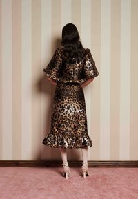 Φόρεμα λεοπάρ με ρυακιές στο τελείωμα, φουσκωτά μανίκια και εφαρμοστή μέση. Το μοντέλο φορά μπεζ ψηλοτάκουνα πέδιλα και στέκεται απέναντι από οριζόντιους τοίχους.