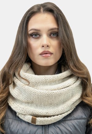Chunky knit cream infinity scarf wrapped around the neck, featuring a textured pattern and a brown leather tag, worn with a dark jacket.