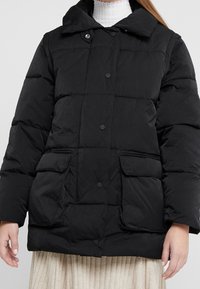 Abrigo acolchado negro con cuello alto, que presenta botones a presión, dos bolsillos frontales y una textura acolchada. Adecuado para el clima frío.