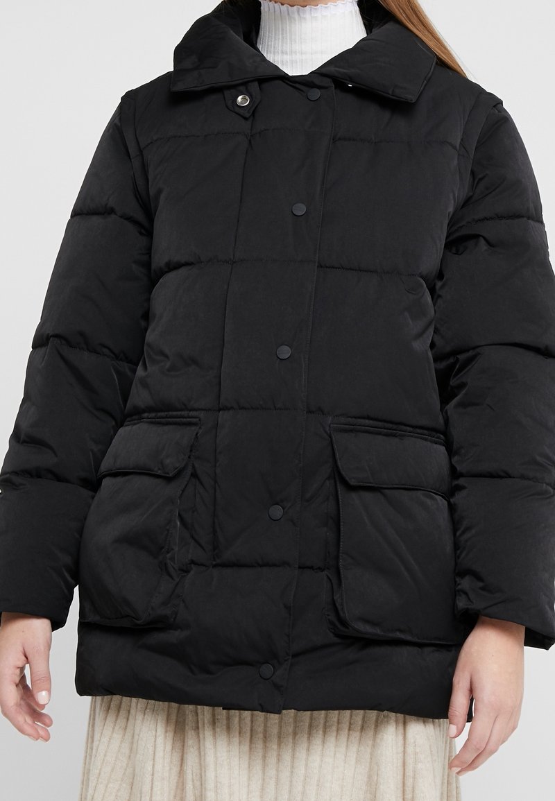 Abrigo acolchado negro con cuello alto, que presenta botones a presión, dos bolsillos frontales y una textura acolchada. Adecuado para el clima frío.