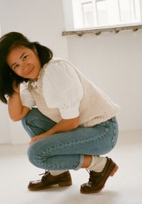 Gilet beige in maglia sopra una camicia bianca, jeans azzurri chiari e scarpe brown con lacci e suola spessa; persona in posa in posizione accovacciata.