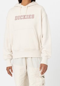 Krämfärgad huvtröja med en framficka, ribbade ärmslut och justerbar huva. "DICKIES"-logotyp i röd på bröstet, avslappnad passform.