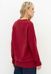 Eine Frau mit blonden Haaren, die einen lockeren roten Pullover und dunkle blaue Hosen trägt, steht seitlich vor einem schlichten weißen Hintergrund.