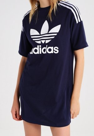 Personne portant une robe t-shirt Adidas bleu marine avec logo trèfle blanc et trois bandes sur les épaules, debout devant un fond uni.