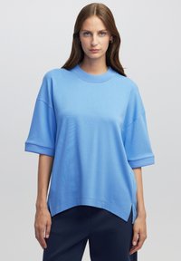 Světle modrý top s krátkým rukávem a kulatým výstřihem. Má uvolněný střih, žebrovaný výstřih a manžety, a boční rozparky na spodním lemu.
