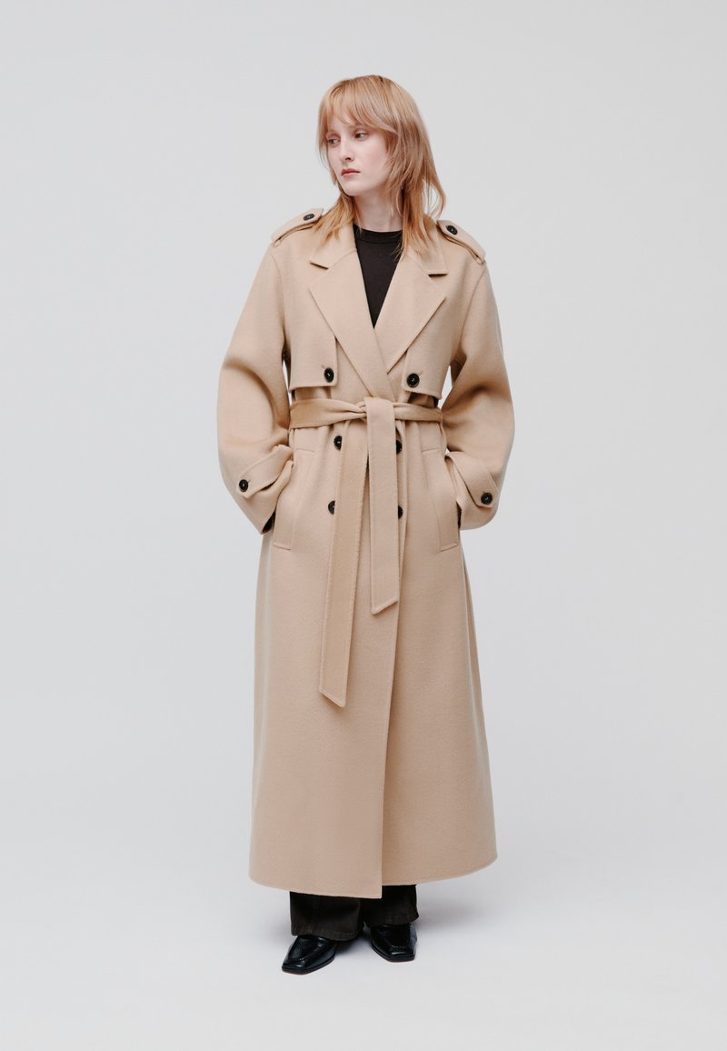Trench coat di lana beige con ampi risvolti, design doppiopetto, maniche larghe e cintura coordinata. Presenta bottoni neri e tasche laterali.