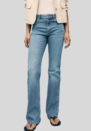 Person, die hellblaue Bootcut-Jeans, eine beigefarbene Jacke und schwarze Sandalen mit Nieten trägt, steht vor einem grauen Hintergrund.