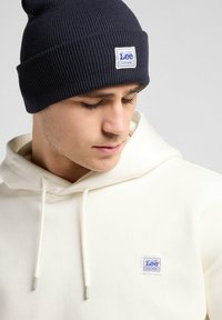 Navy ribbed beanie and cream hooded sweatshirt. Beanie features small logo patch; sweatshirt has logo patch on the left chest.