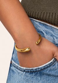 Pulsera de pulsera de metal dorado con una superficie suave y reflectante y extremos redondeados, llevada en una muñeca por encima de un bolsillo de mezclilla.
