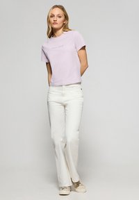 Lichtpaarse T-shirt met korte mouwen met de tekst "Pele Club". Gecombineerd met witte rechte jeans en beige sneakers, neutrale achtergrond.