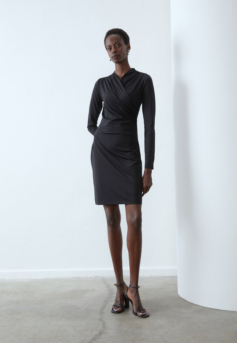 Abito lungo nero a maniche lunghe con silhouette aderente, design avvolgente e texture liscia. Abbinato a sandali neri con tacchi alti e fascette.
