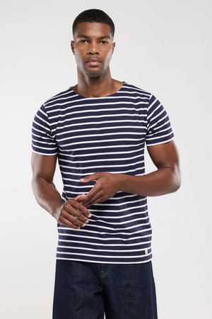 Jeune homme portant un t-shirt rayé bleu marine et blanc, ajustant ses doigts, se tenant devant un fond blanc uni.