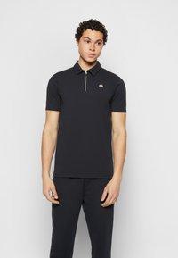 Čierna polo košeľa s golierom na zips, s malým logom na ľavom prsníku. Materiál vyzerá hladko, má krátke rukávy a voľný strih.