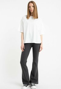 Młoda kobieta stoi w luźnej białej koszulce, dopasowanych ciemnoszarych spodniach typu dzwony i czarno-białych sneakersach na białym tle.