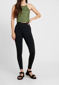Zelený leopardí vzorovaný crop top, černé legíny a černé sandály. Top má tenká ramínka a volný střih; legíny jsou těsně padnoucí.