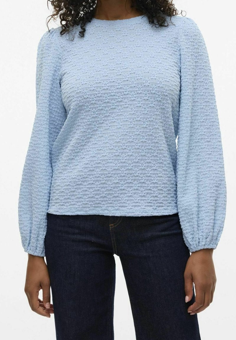 Light blue textured blouse with long puffed sleeves and a round neckline, paired with dark denim jeans.