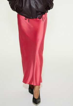 Personne portant une jupe en satin rouge brillant associée à une veste en cuir noire et des escarpins noirs à bout pointu, debout devant un fond uni.