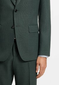 Mann trägt einen dunkelgrünen, strukturierten Anzug mit zwei schwarzen Knöpfen, Pattentasche und sichtbarem hellblauen Hemdsärmel an der rechten Handseite.